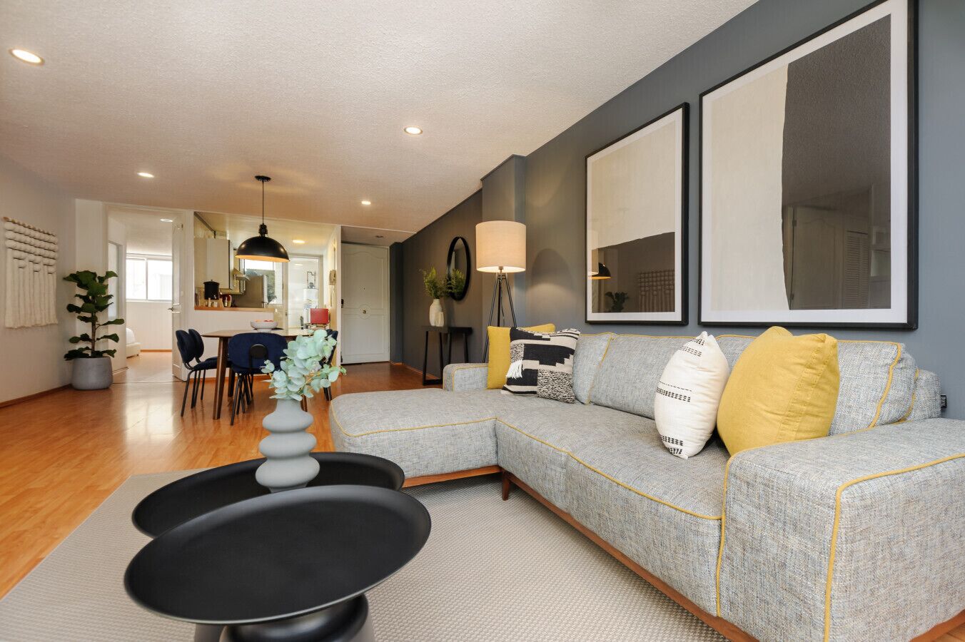 Een moderne woonkamer met een grijze bank, gele kussens en een zwarte salontafel. Op de achtergrond is een eethoek met een witte tafel en stoelen.