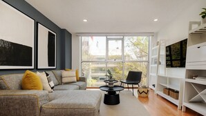 Un salon moderne avec un canapé gris, une table basse noire et une grande fenêtre donnant sur des immeubles et des arbres.