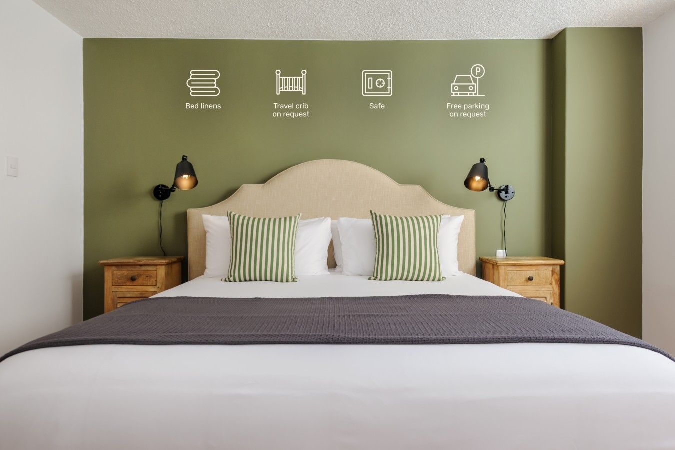 A hotel room with a bed, two bedside tables, and a wall with icons indicating bed linens, a travel crib on request, safety, and free parking on request.