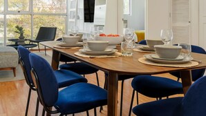Un espace repas moderne avec une table en bois dressée pour quatre personnes, entourée de chaises recouvertes d’un tissu bleu. Un grand luminaire suspendu noir est placé au-dessus de la table.