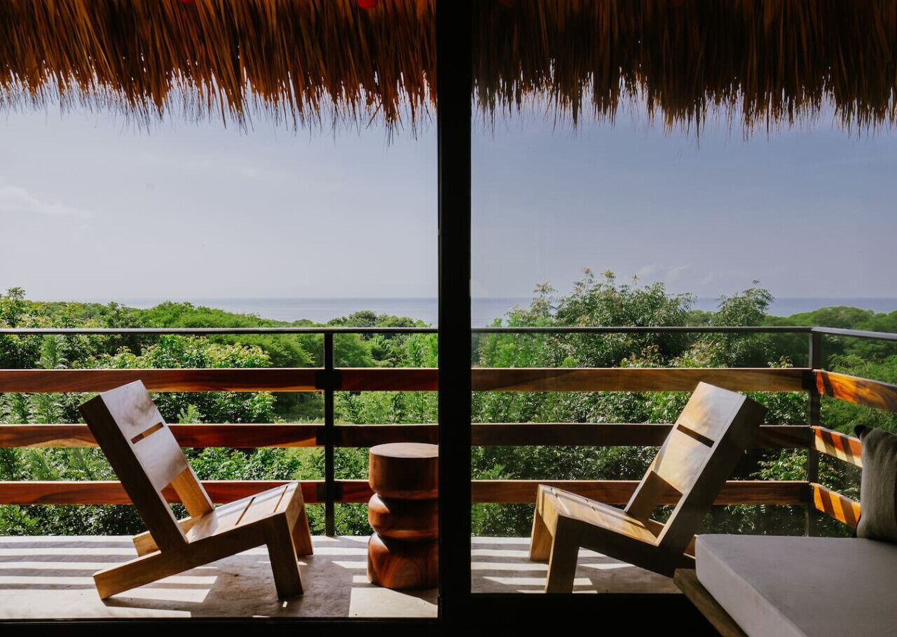 Un balcon doté de deux fauteuils de jardin en bois et d’une petite table ronde, offrant une vue sur un paysage verdoyant et luxuriant ainsi que sur la mer.