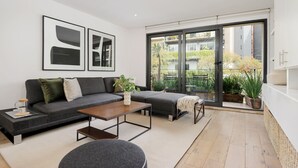 Ein modernes Wohnzimmer mit einem grauen Sofa, einem Couchtisch und einem runden Hocker. An der Wand hängen zwei gerahmte abstrakte Gemälde, und eine Schiebetür führt auf einen Balkon mit Topfpflanzen.