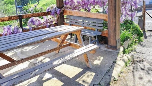 Una pérgola de fusta amb una cadira i taula, que dona a un jardí ple de flors morades i vista cap a cases a la distància.