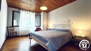 Un dormitori amb sostre de fusta, un llit amb estructura metàl·lica, una cadira, una taula de nit amb una làmpada, i una finestra amb cortines transparents.