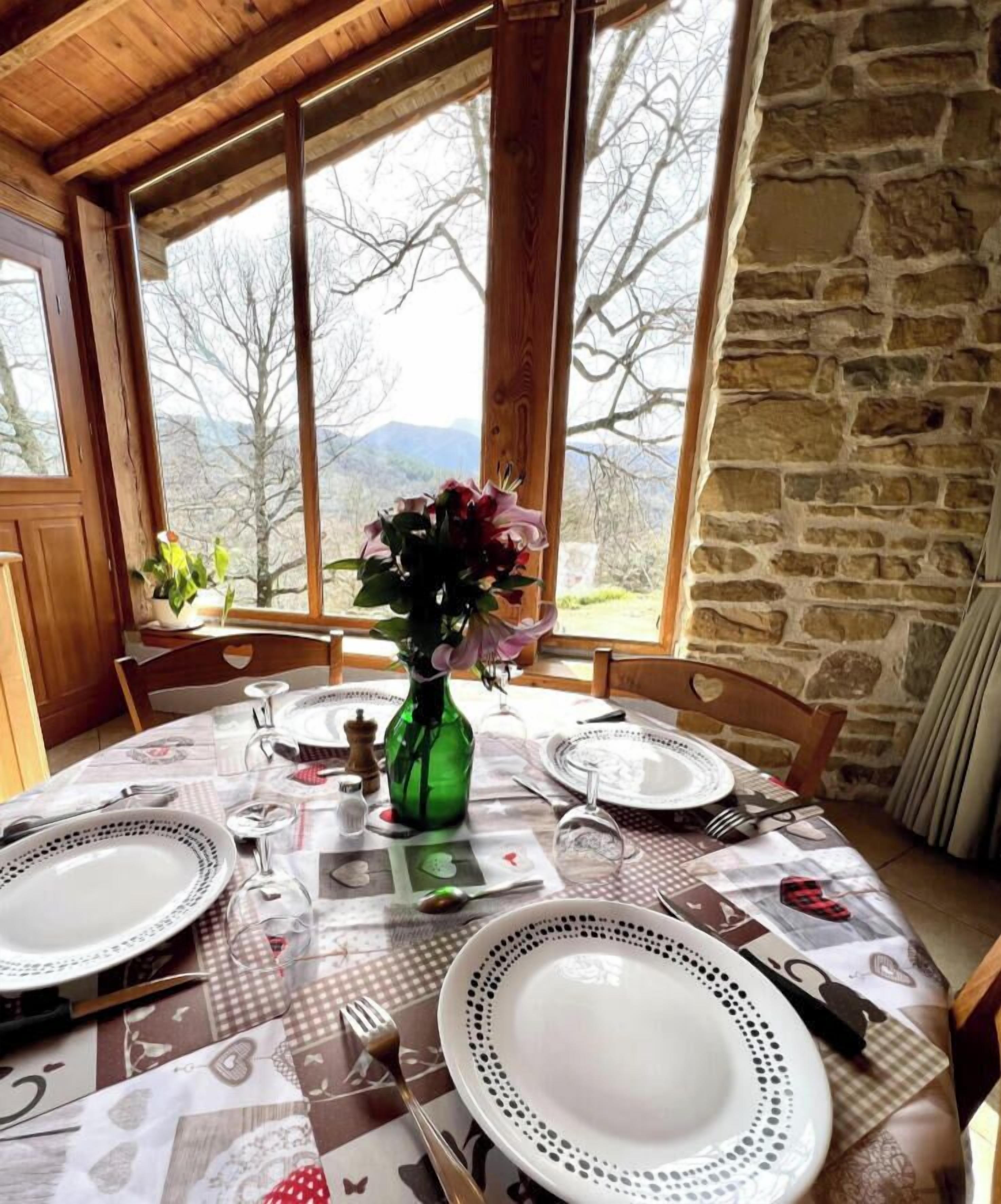 A dining table set with plates, cutlery, and a green bottle, in a room with stone walls and large windows.