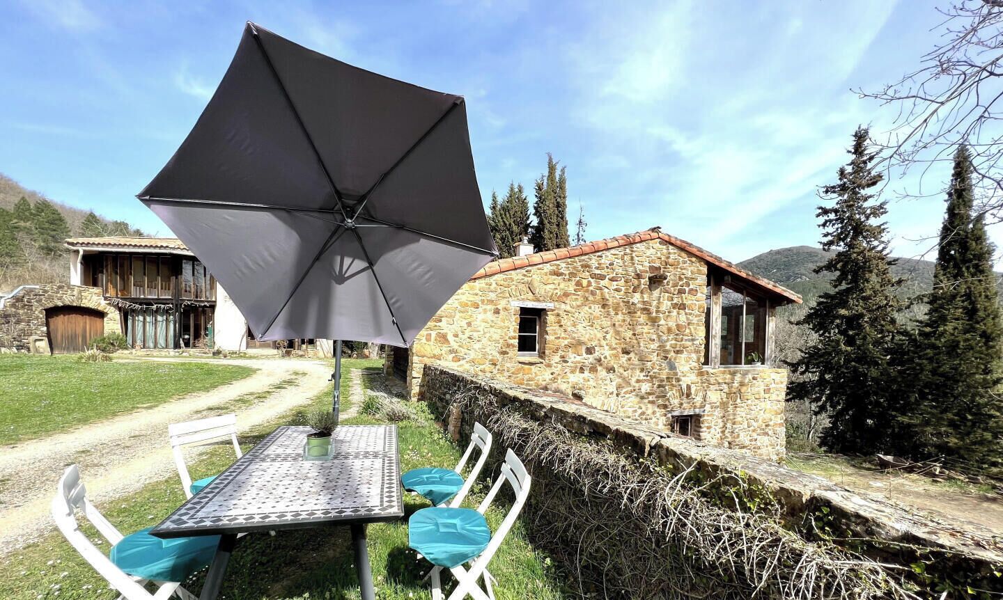 A stone building with a thatched roof, a table with chairs, and a large umbrella.