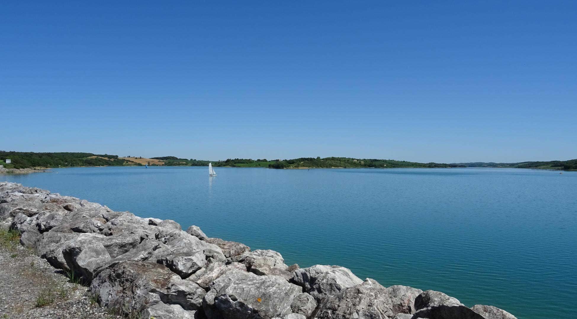 A calm lake with a sailboat, rocky shoreline, and distant green hills.