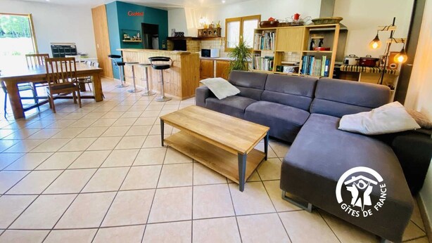 A modern living room with a sofa, a coffee table, and a dining area with chairs and a table. There is a kitchen area with a bar and a bookshelf filled with books. - Les Jardins du Plateau (ESPEZEL)