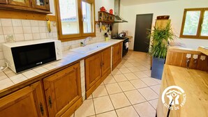 A kitchen with wooden cabinets, a microwave, a sink, and a stove. There is a window with a view of greenery outside. - Les Jardins du Plateau (ESPEZEL)
