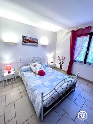 Ein Schlafzimmer mit einem Metallbettgestell, einem Nachttisch mit Lampe, einer Wandleuchte, einem Fenster mit Vorhängen und einem Gemälde an der Wand.