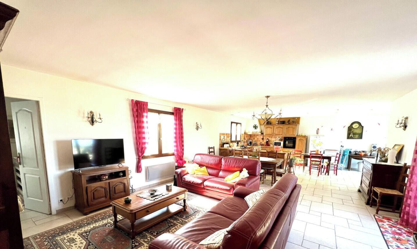 A living room with a sofa, a coffee table, and a television. There is a kitchen in the background with wooden cabinets and a dining area.