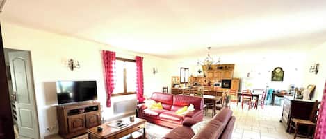 A living room with a sofa, a coffee table, and a television. There is a kitchen in the background with wooden cabinets and a dining area.