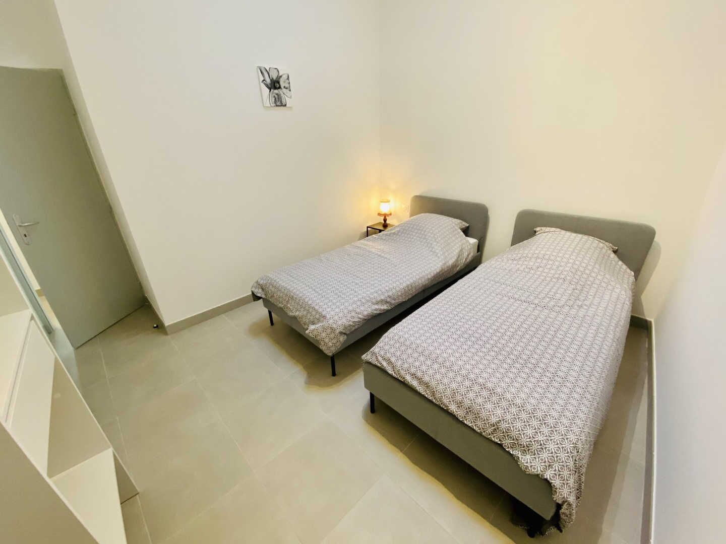 A small, clean room with two beds, a nightstand with a lamp, and a door.