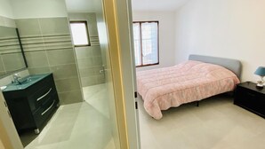 Ein Schlafzimmer mit Bett, Nachttisch, Lampe, Fenster und einem Badezimmer mit Waschbecken und Spiegel.