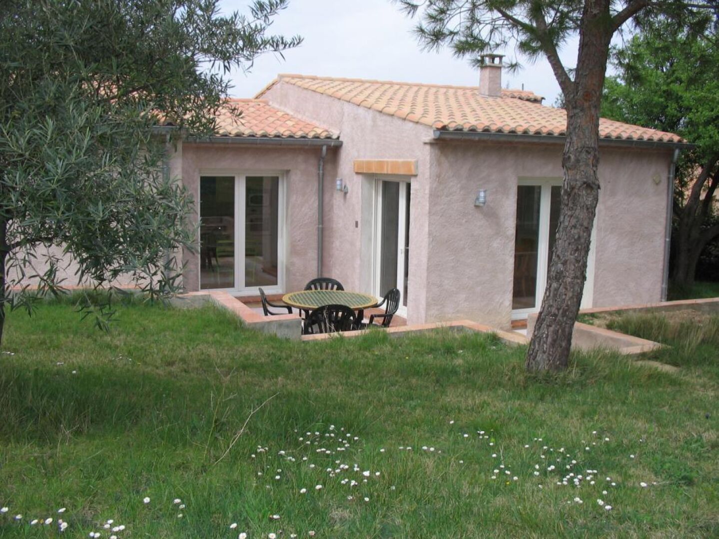 Ein eingeschossiges Haus mit Ziegeldach, einer Terrasse mit Tisch und Stühlen sowie einem Rasen mit weißen Blüten.