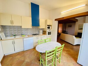 A compact kitchen with white cabinets, a blue accent wall, a dining table with chairs, and a visible living area with a sofa and TV. - Gite des Pesquis - BAGES (BAGES)