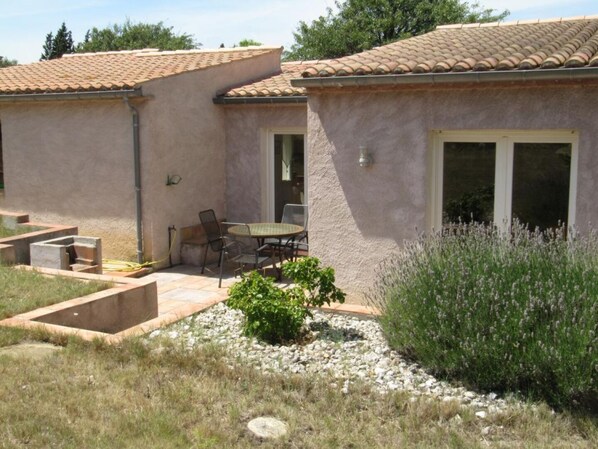 A building with a tiled roof, a patio area with a table and chairs, and a garden with plants and a gravel path. - Gite des Pesquis - BAGES (BAGES)