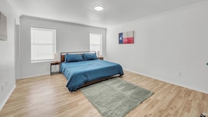 Une chambre à coucher comprenant un lit, deux tables de chevet, une commode, une fenêtre avec des stores, un luminaire au plafond et une œuvre d’art accrochée au mur.
