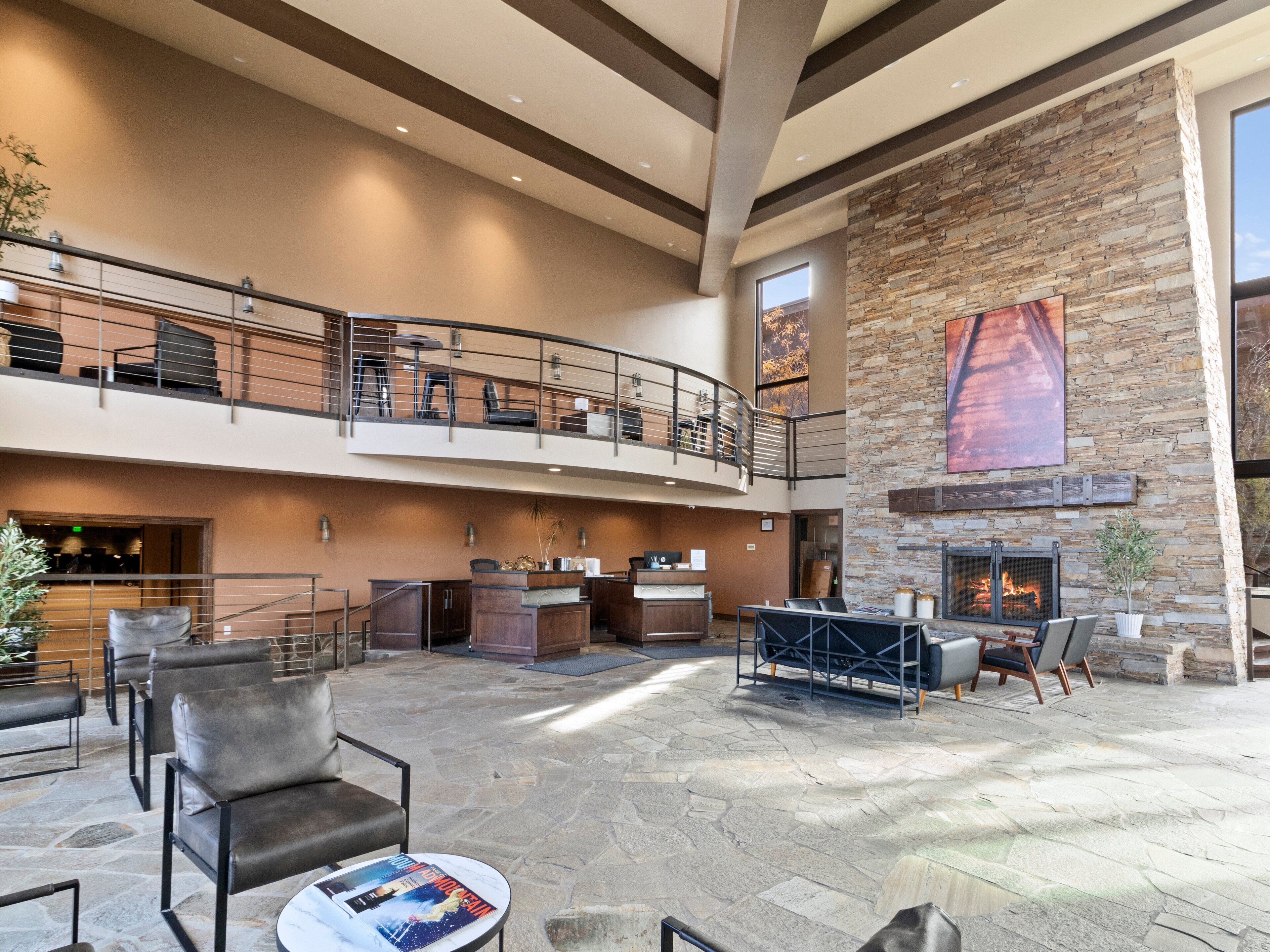 Un hall d’entrée spacieux avec un sol en pierre, une cheminée, un comptoir de réception et un balcon au deuxième étage, aménagé avec des sièges.