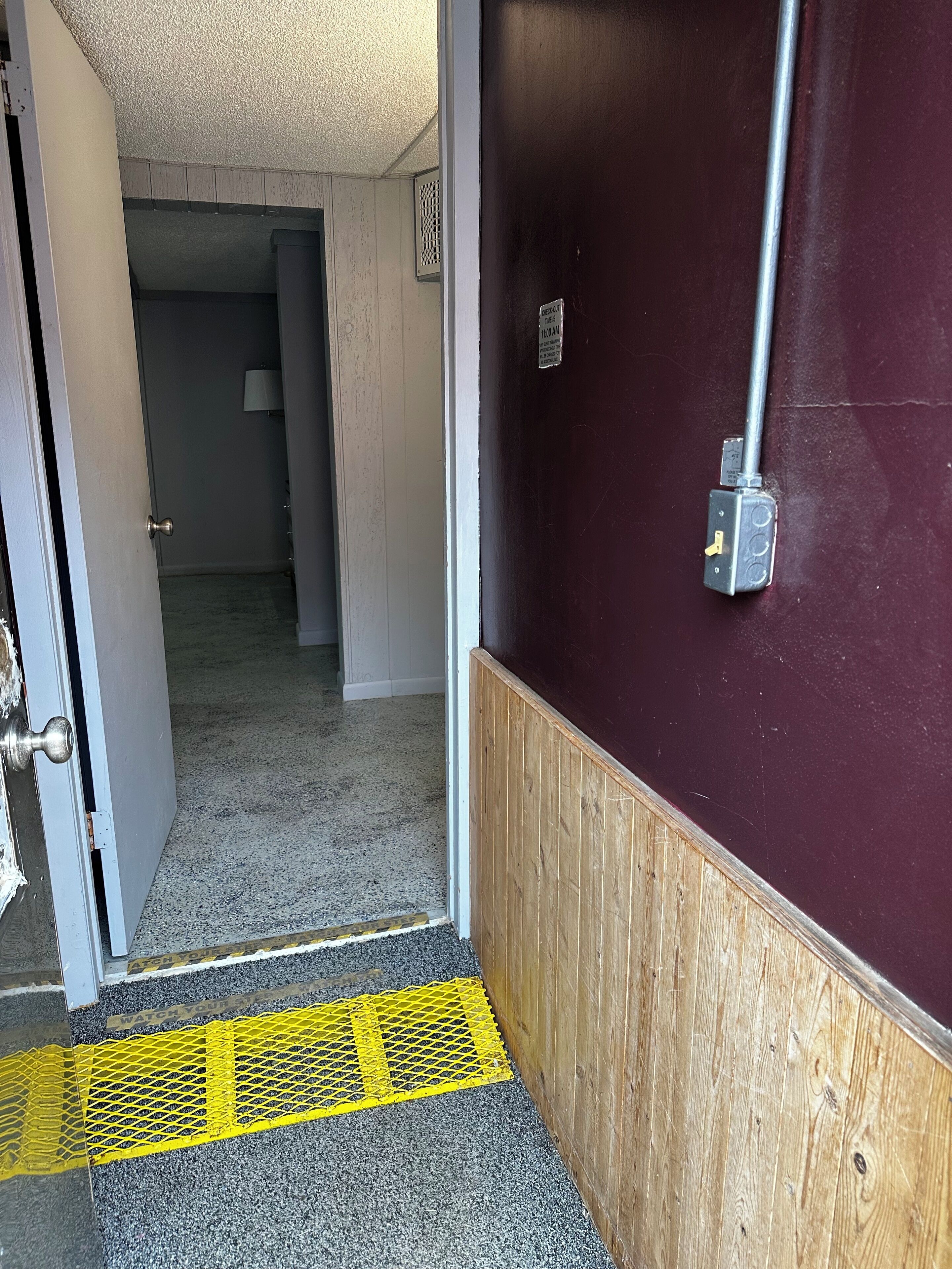 L’entrée d’un hôtel, avec un panneau en bois, une porte en métal et un chemin tactile jaune.