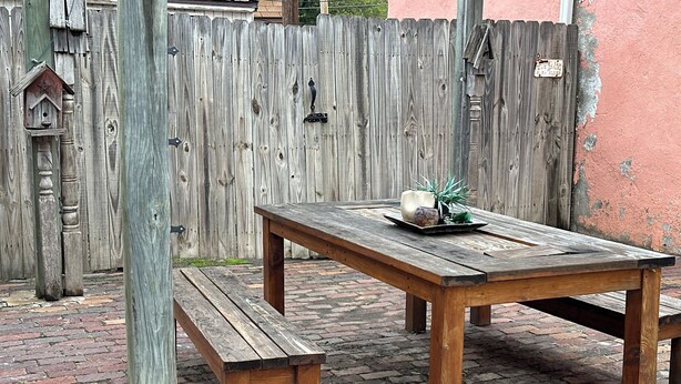 Une table de pique-nique en bois avec des bancs, un nichoir décoratif et une plante en pot sur une terrasse en briques.