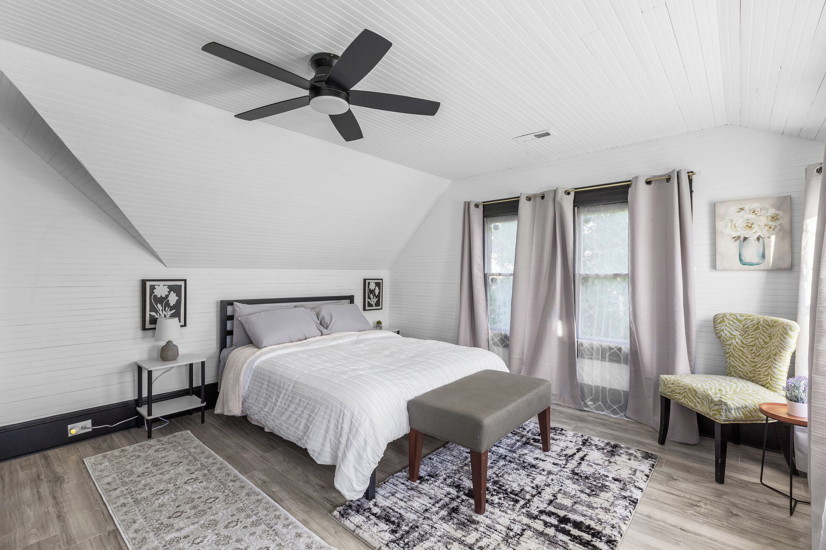 Une chambre à coucher avec un lit, un ventilateur de plafond, une fenêtre avec des rideaux, une chaise et un tapis.