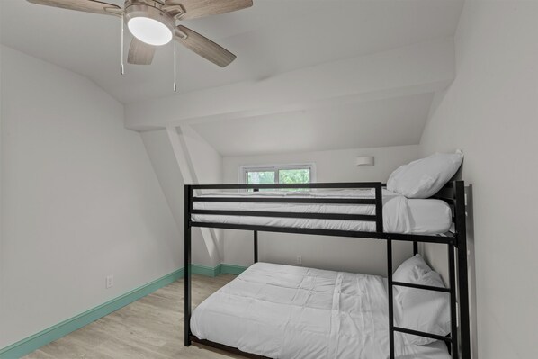 A bunk bed room with a ceiling fan, white walls, and a window with a view of greenery.