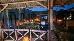 Estructura de bambú con techo de paja, rodeada de plantas de interior e iluminada por luces cálidas.