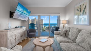Ein Wohnzimmer mit Fernseher, einer Couch, einem Sessel, einem Couchtisch und einem Balkon mit Meerblick.