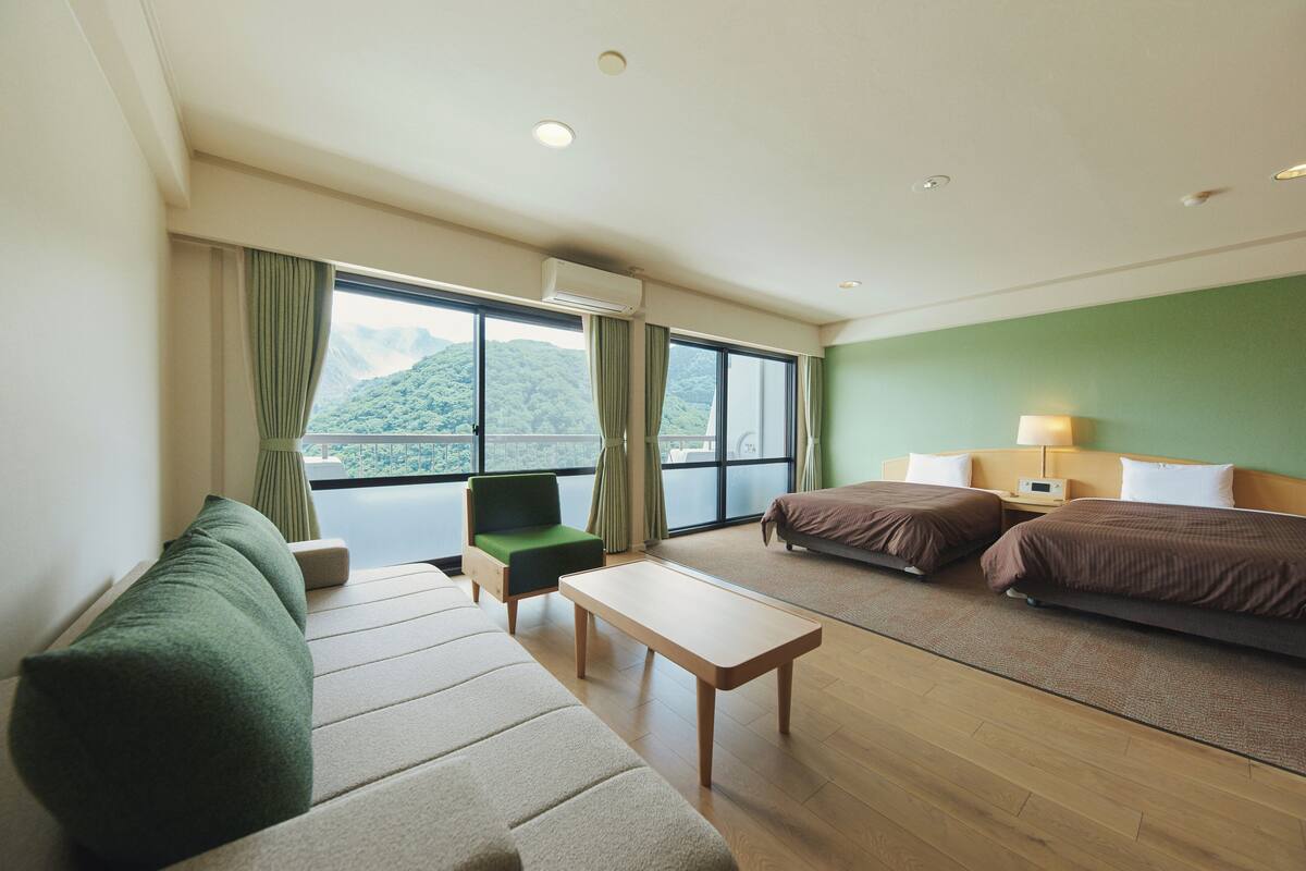 Une chambre d’hôtel avec un canapé, deux lits, une petite table et une vue sur les montagnes grâce à de grandes fenêtres.