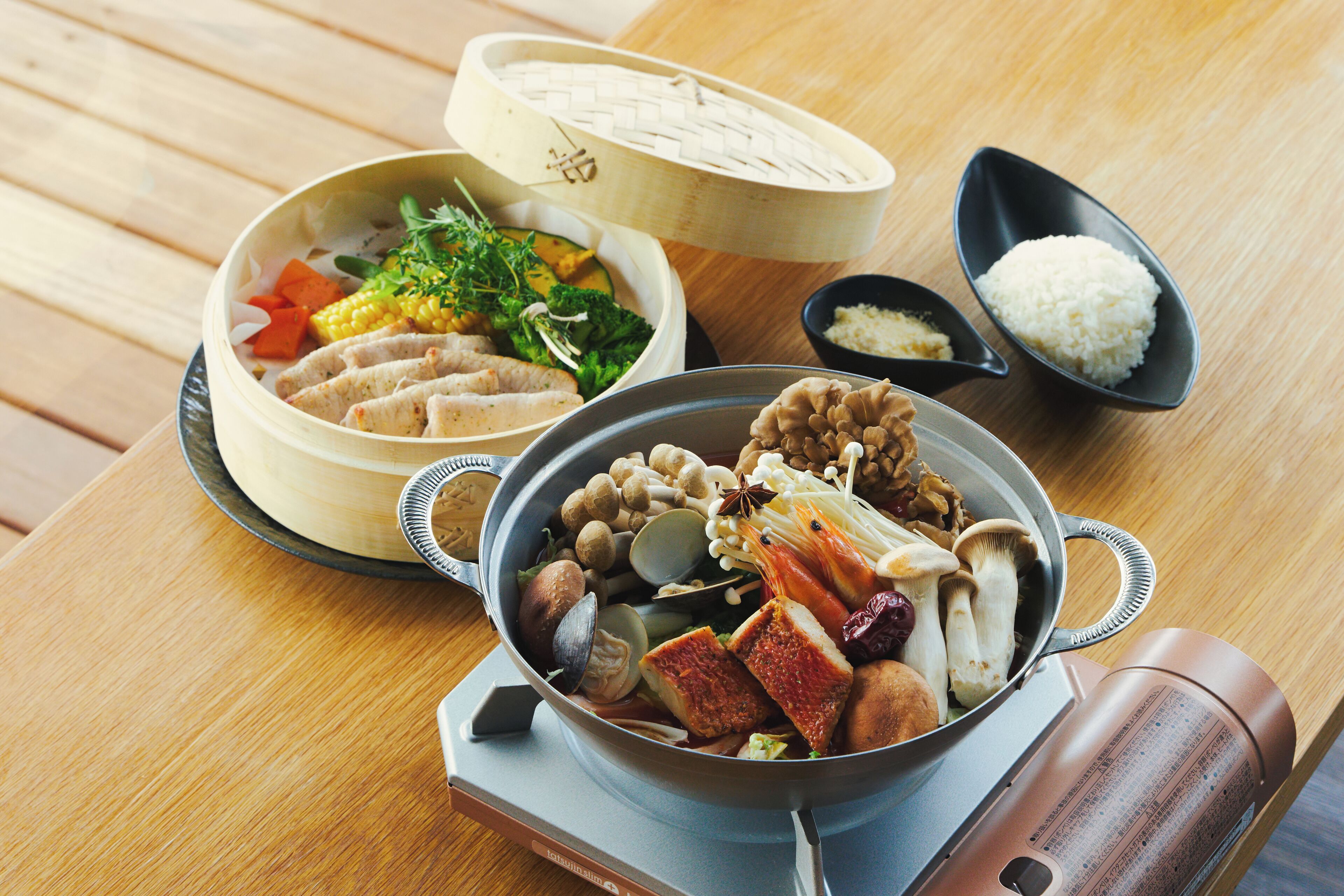 A wooden table with a meal consisting of a bowl of soup, a bowl of rice, and a pot of assorted vegetables and mushrooms.