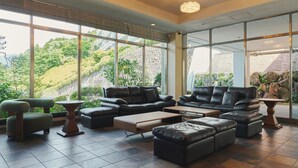 Ein großzügiger Innenbereich mit Ledersofas und Poufs, die um einen zentralen Couchtisch angeordnet sind, große Fenster mit Blick auf eine Hügelkette und eine sichtbare Steinwand durch das Glas hindurch.