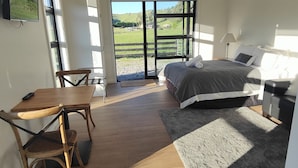 A bedroom with a bed, a desk, and chairs, a TV, and a large window with a view of a green landscape. - Katelyn Studio  - Rural Couples Retreat (Kinloch)