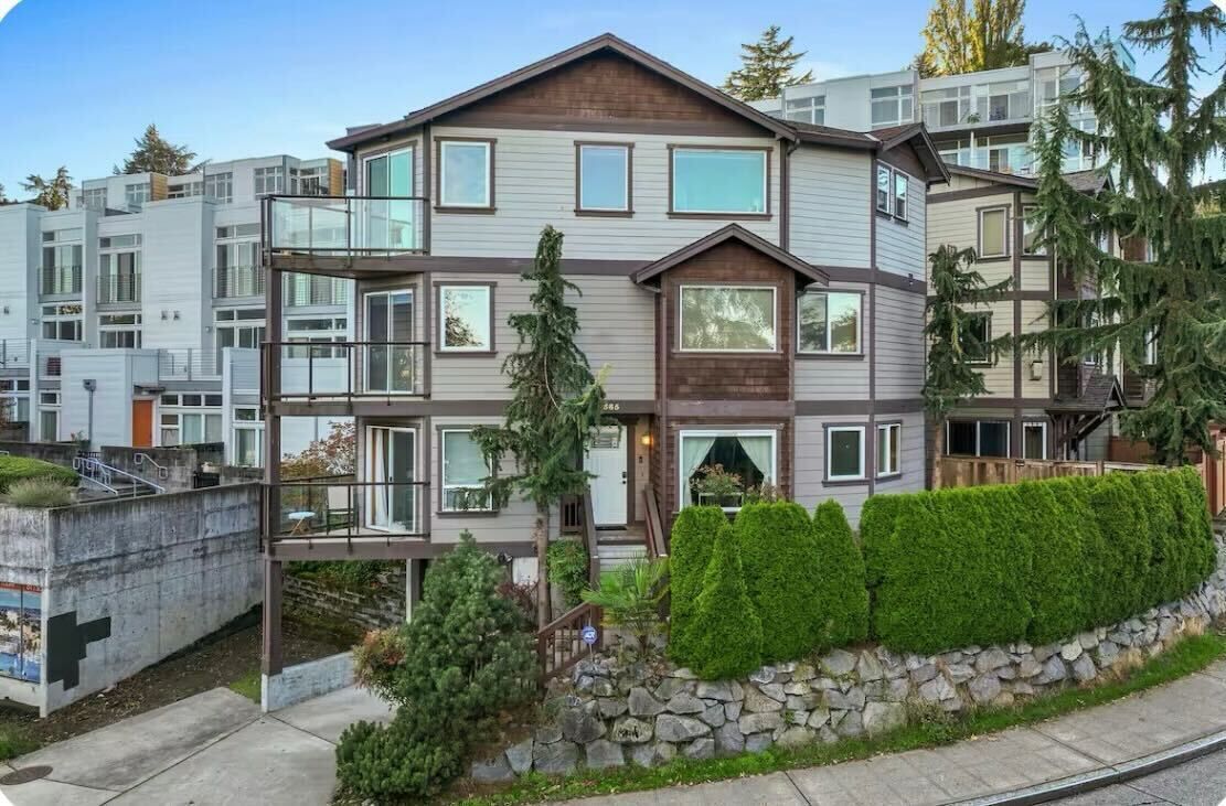 A multi-story residential building with a modern design, featuring balconies, large windows, and a stone retaining wall with greenery.