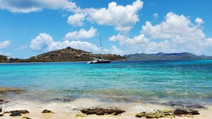 A coastal scene with clear turquoise waters, a sandy beach with rocks, and a sailboat anchored near a hill with buildings. - Oceanfront Ritz-Carlton Condo 2BR 2.5BA, Full Resort Amenities! (St Thomas)