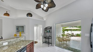 A kitchen with granite countertops, a dishwasher, and a ceiling fan. There is a dining area with outdoor furniture visible through a glass door. - New! Salty Steps - Naples Getaway with Hot Tub & Outdoor Kitchen (Naples)