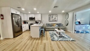 A modern kitchen with white cabinets, a stainless steel refrigerator, and a microwave. There's a gray sofa with patterned cushions in the living area, and a small white coffee table. - NEW!! Rare 5 bedroom house w FREE HEATED POOL & HOT TUB 9 minutes to the beach!! (Seminole)