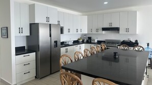 A modern kitchen with white cabinets, a stainless steel refrigerator, a black countertop, and a dining area with wooden chairs. - Luxury Beach house , infinity, pool, breathtaking view, Surf city,Sunset Park. (La Libertad)