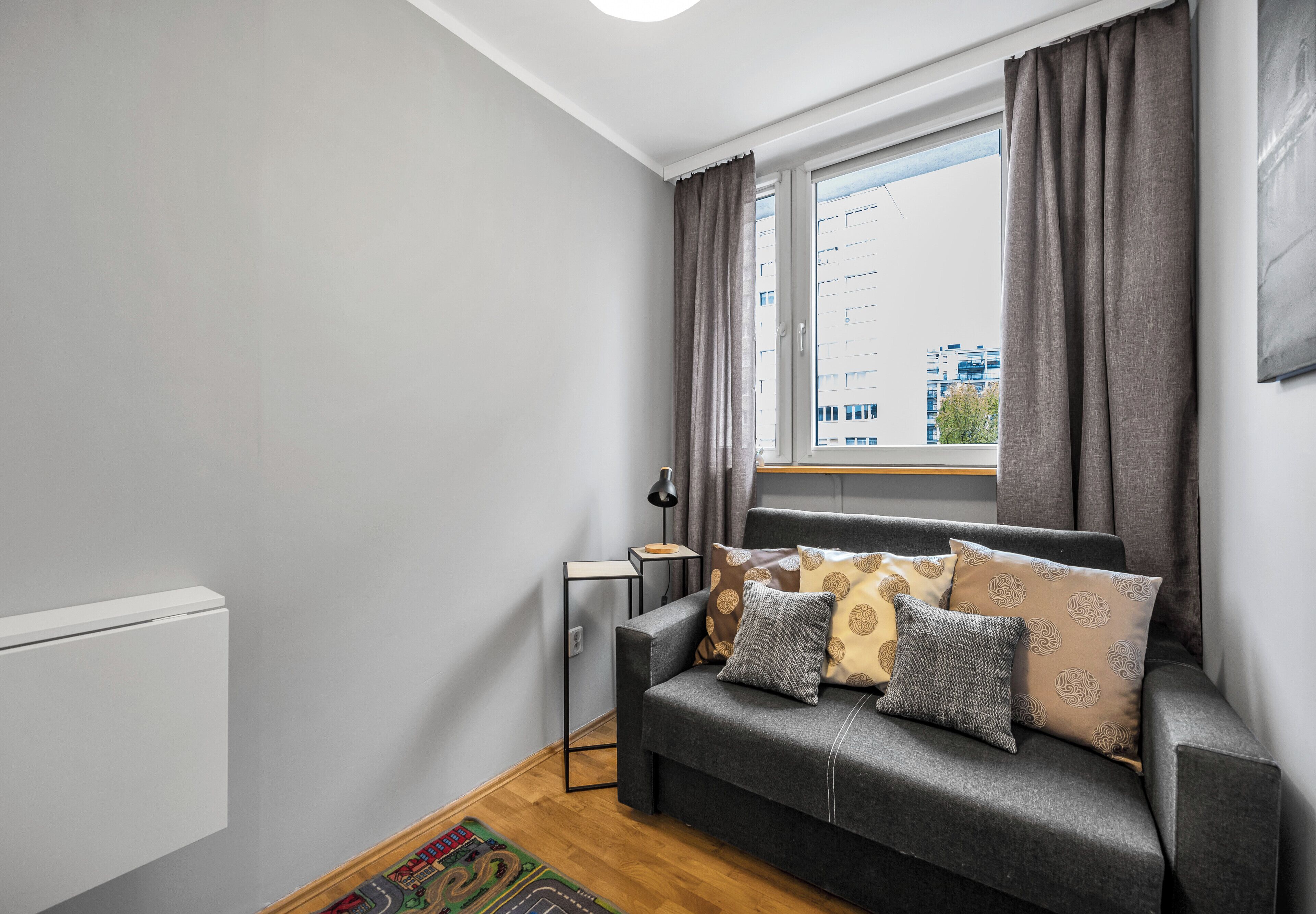 A compact living space with a grey sofa, patterned cushions, a small side table, and a window with curtains.