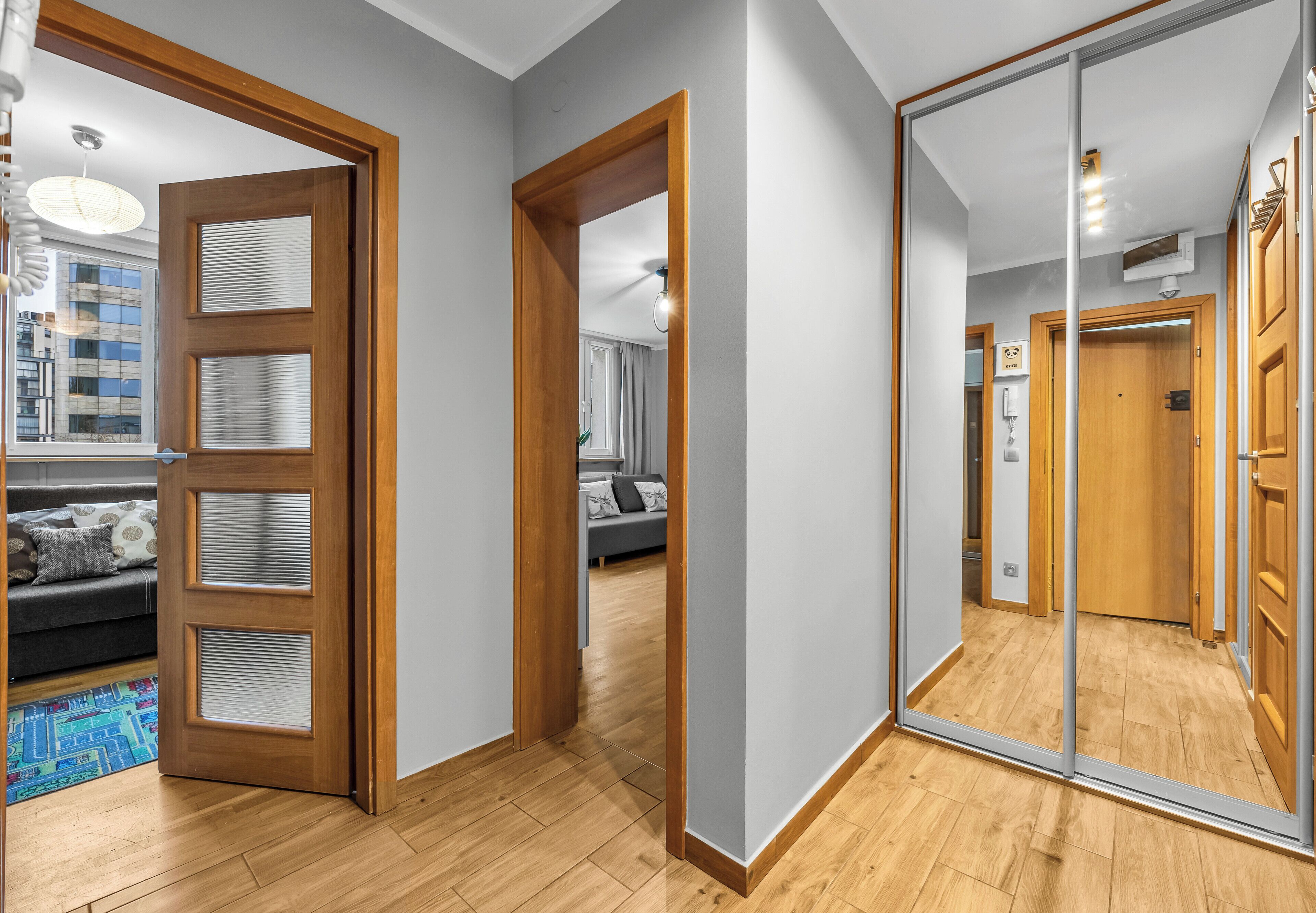 Un pasillo con puerta de madera, un espejo grande y vista a un área de estar con sofá y alfombra.