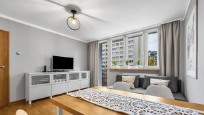 Een moderne woonkamer met een houten eettafel, een televisie op een witte kast, een bank met kussens en een groot raam met gordijnen.