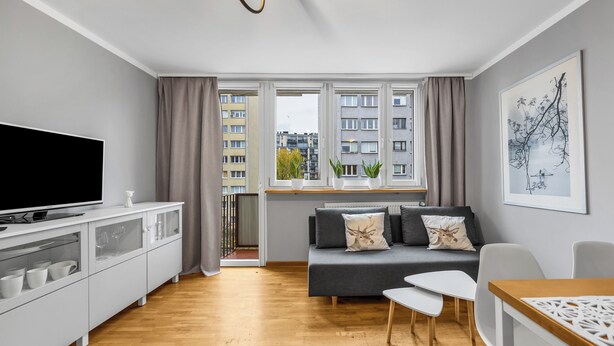 Een moderne woonkamer met een flatscreen-tv, een bank met kussens, een eettafel en een raam met gordijnen.