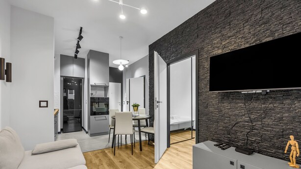 A modern living room with a sofa, a dining area with chairs, a kitchen in the background, and a TV mounted on a stone-textured wall. - Kłobucka Deluxe Apartment (Warszawa)