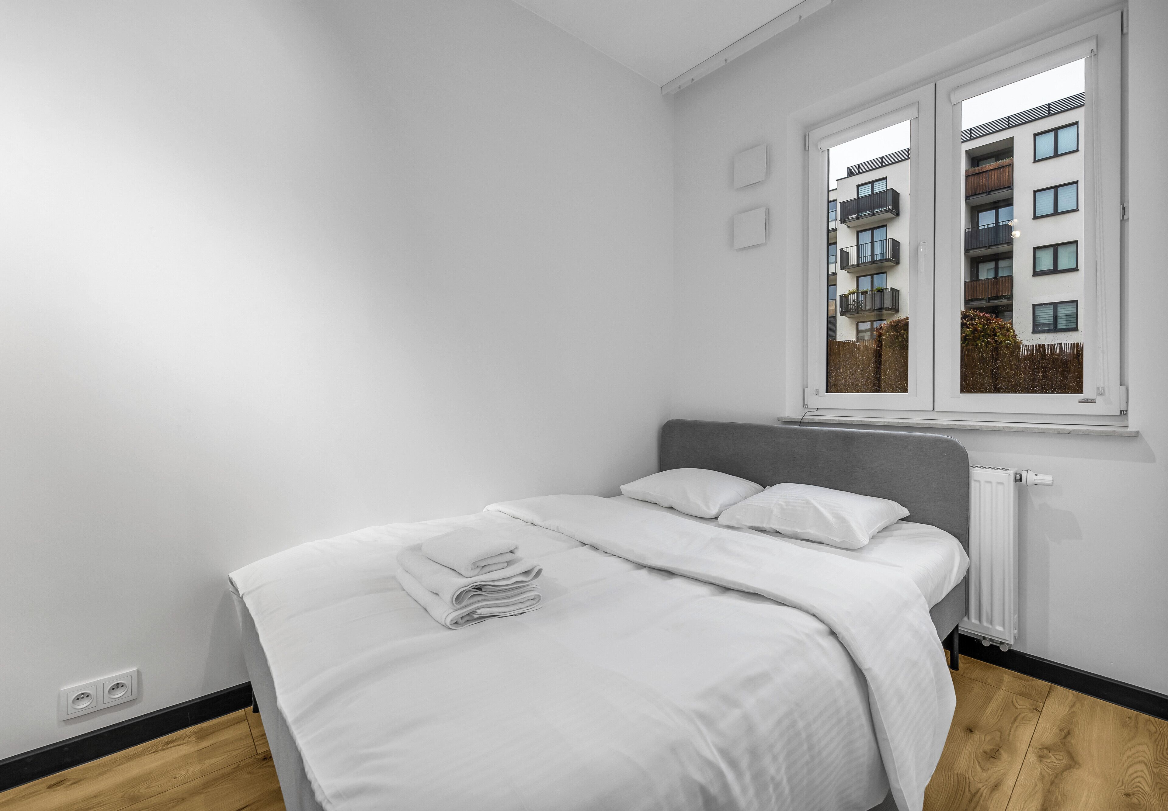 Un dormitorio minimalista con cama, ropa de cama blanca, un radiador y una ventana con vista a un edificio.