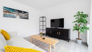 Ein modernes Wohnzimmer mit einer Couch, einem Couchtisch, einem Fernseher auf einem schwarzen Schrank, einer stehenden Stehlampe und einer Topfpflanze.