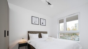 Une chambre moderne avec un grand lit, une table de chevet, une fenêtre donnant sur la ville et deux œuvres d’art encadrées accrochées au mur.