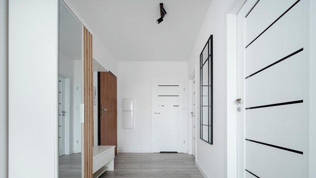 Un couloir aux murs blancs, une porte en bois, une armoire blanche et une porte blanche ornée de lignes horizontales noires.