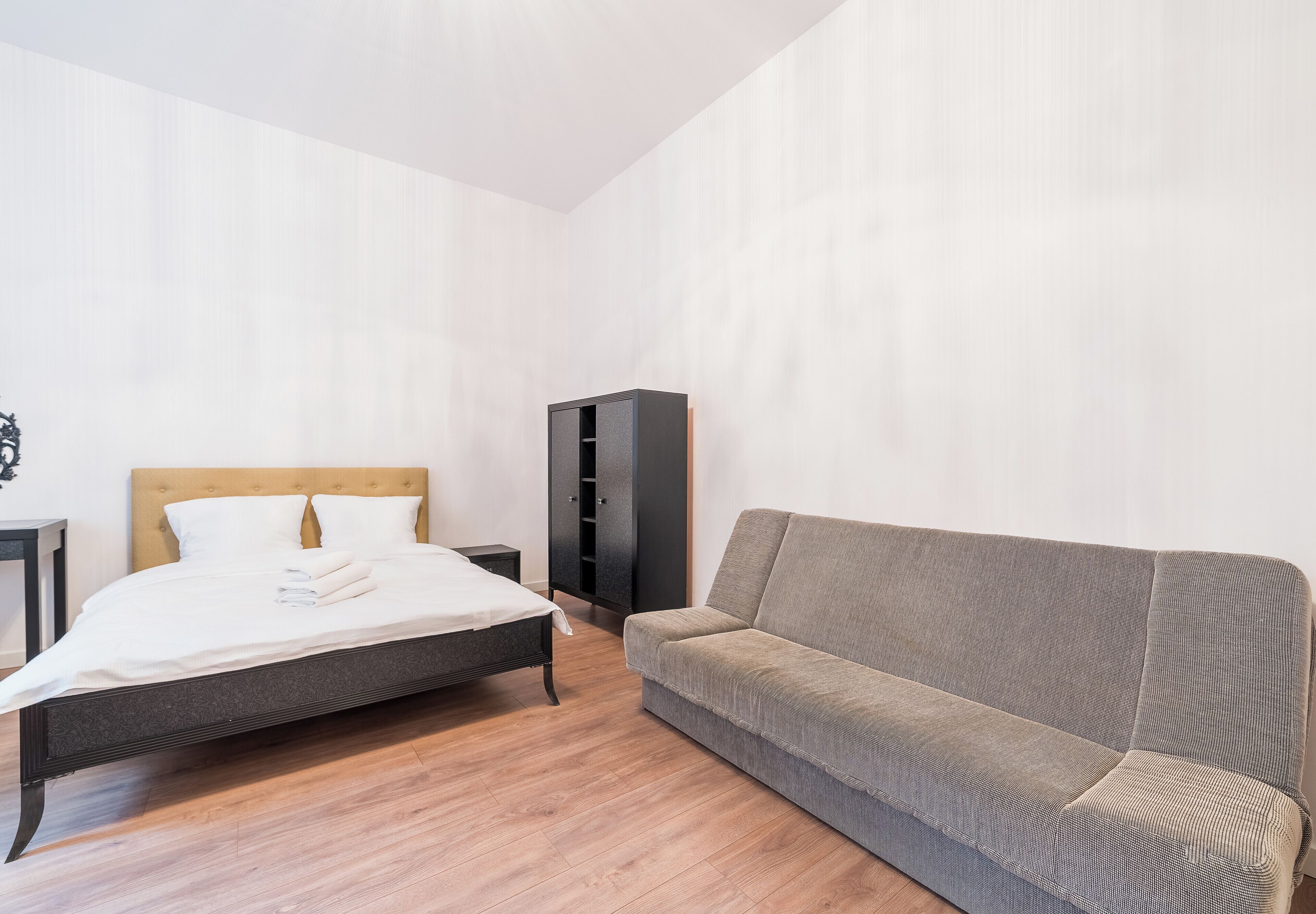 A bedroom with a bed, a grey sofa, a wardrobe, and a wooden floor.