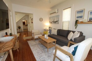 Ein Wohnzimmer mit Sofa, Couchtisch und Sesseln, im Hintergrund ein Küchenbereich und eine Treppe, die zu einer weiteren Ebene führt.