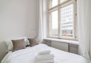 A neatly made bed with white linens and a folded towel, a window with white curtains, and a view of a building outside. - Heart of Poznań Studio #1 (Poznań)